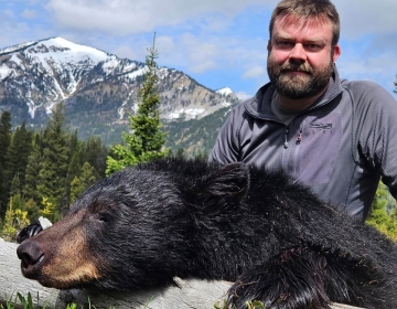 A nonresident black bear hunter poses with his trophy over a log with a snow covered mountain range in the background.